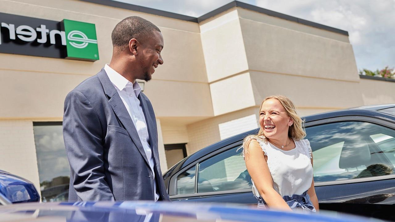 一个年轻的男人和一个年轻的女人，在一个阳光明媚的日子里，微笑着谈论着企业租车公司的停车场.